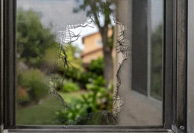 Window screen with a large tear in the mesh, in an aluminum frame on a stucco house